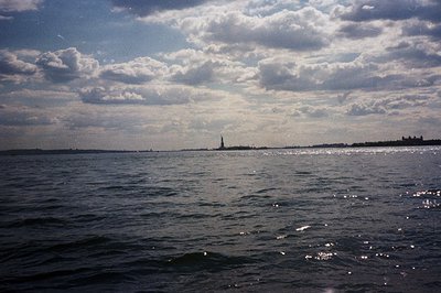 A long, wide shot captures the Statue of Liberty across a shimmering body of water, likely New York Harbor. The sky is overca...