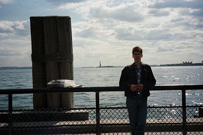 A young man stands near a pier, gazing toward the Statue of Liberty and the Manhattan skyline. He wears a dark blazer and jea...