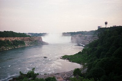 A wide, sweeping view captures Niagara Falls, partially obscured by mist and spray. Lush green vegetation clings to the rocky...