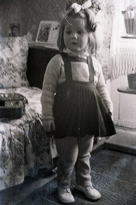 A young girl stands in a domestic interior, wearing a cable-knit sweater, dark overalls, and a ribboned headband. The room fe...