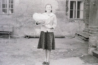 A woman stands in a courtyard, cradling a baby. She wears a patterned skirt and a light jacket. The architecture suggests a E...