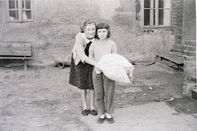 Two girls stand in a courtyard, one embracing the other who carries a large cloth bag. Brick buildings and a wooden bench for...