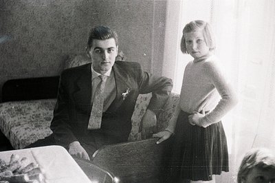 Man in suit & tie sits formally; a young girl stands beside him, hands on hips. Interior features patterned wallpaper & a bed...