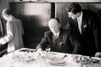 A formal dining scene: a seated man in a suit observes a lavish table laden with food and glassware. A younger man in a suit ...