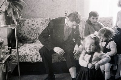 Man in a suit kneels to interact with two young girls. Floral patterned sofa, potted plant, and vintage furniture suggest a m...