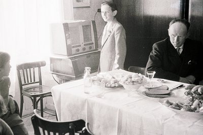 A family gathers around a table laden with food, featuring a vintage radio set prominently displayed. A young person stands n...