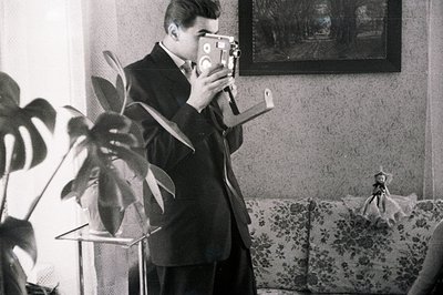 A sharply dressed man operates a vintage 8mm movie camera, likely filming within a domestic setting. The scene includes a pot...