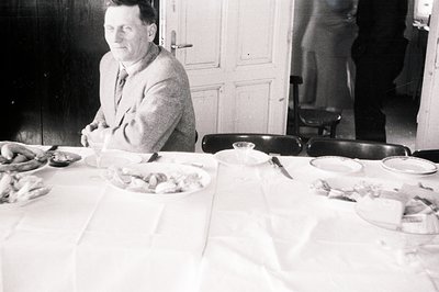 A seated man in a tweed suit observes a lavishly set table, possibly during a celebration. Numerous dishes of food—meats, che...