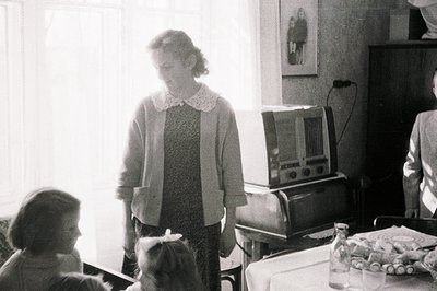 A seated young girl and a woman in a patterned dress and cardigan stand in a domestic interior. A large vintage radio dominat...