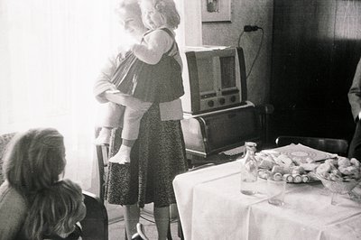 A woman in a patterned dress holds a small child on her hips, standing near a large, vintage radio console. A table with glas...