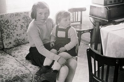 Two young girls, likely sisters, posed indoors. The older girl wears a pleated skirt & sweater; the younger sits on her lap i...