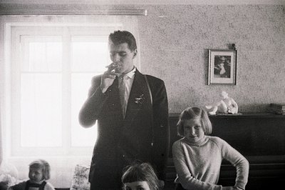 Formal portrait of a young man in a suit smoking, flanked by two girls seated near a piano. Wallpapered interior, likely a fa...