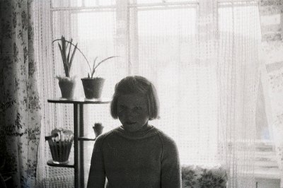 Young girl stands near a window, framed by sheer curtains & potted plants on a decorative shelving unit. Likely mid-20th cent...