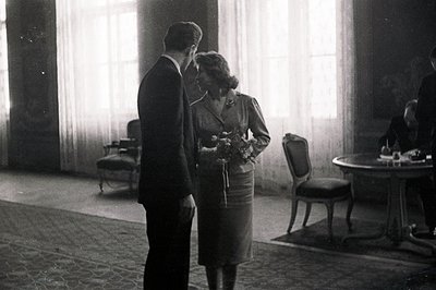 A formally dressed couple stands facing each other in a grand, light-filled room. The woman holds a bouquet of flowers. Visib...