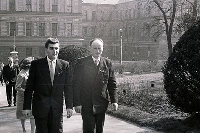 Two formally attired men walk along a paved path lined with manicured shrubs, a grand building visible in the blurred backgro...