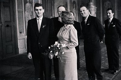 A young bride in a pale suit carries a bouquet, posed with four formally dressed men, likely groomsmen. The setting appears t...