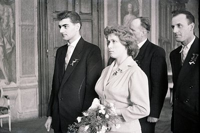 Formal portrait showcases a wedding party. A young couple stands at center, with two older men flanking them. The bride holds...
