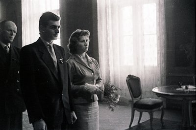 Formal portrait of a couple and a gentleman in a room with large windows. The man wears a suit and tie, she a suit with glove...