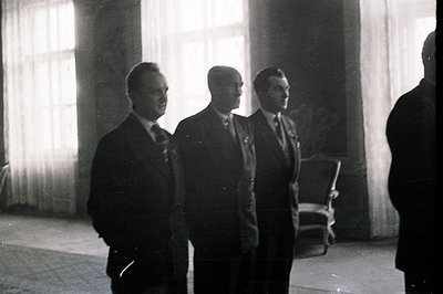 Three formally dressed men stand in a spacious, light-filled room with sheer curtains. The image appears to be a candid momen...