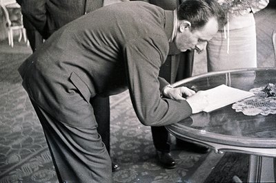 A man in a dark suit bends over a round table, writing on a document placed atop a lace doily. Visible through the table’s gl...
