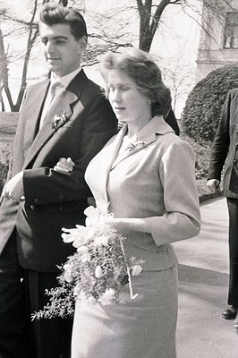 A posed black and white portrait captures a formally dressed couple, likely during a wedding. The woman holds a bouquet of fl...