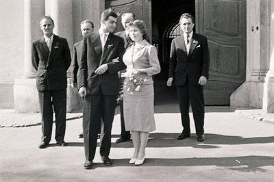 Formal portrait of a wedding party outside a grand building. Bride in a knee-length dress and floral bouquet stands between f...