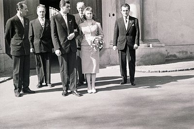 Black & white photo depicts a wedding party exiting a building. Five figures: groom, two groomsmen, the bride, and another ma...