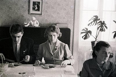 Three formally dressed individuals seated around a covered table. A decorative mantelpiece featuring a statue and framed artw...
