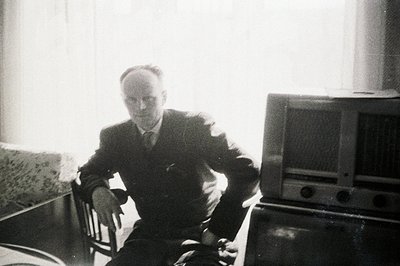 A gentleman in a dark suit and tie sits on a chair facing the camera, positioned next to a vintage radio. The room's light so...
