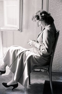 A woman seated in a chair, engrossed in reading a book. She wears a wide-legged suit and dark heels. Natural light illuminate...