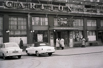 Hotel entrance, likely 1960s, features a prominently signed facade and classic cars parked along the street. Three figures ar...