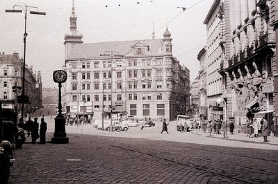 Historic street scene. Ornate, multi-story building with a central clock tower dominates the composition. Cobblestone street ...