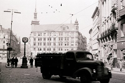 A bustling street scene, likely post-war Europe, features a military transport truck dominating the foreground. Ornate, multi...