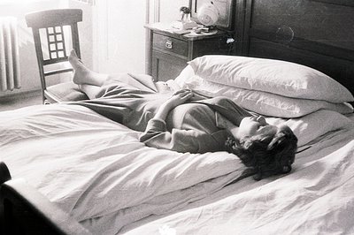A young woman reclines on a heavily draped bed, appearing relaxed or asleep. A wooden chair sits nearby, and a bedside table ...