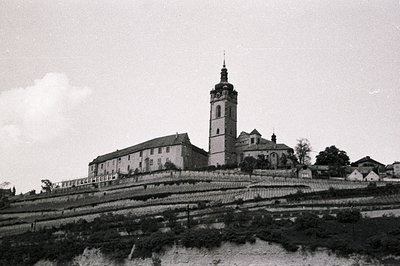 A hillside terraced landscape leads up to a large stone monastery with a prominent clock tower. The building displays multipl...