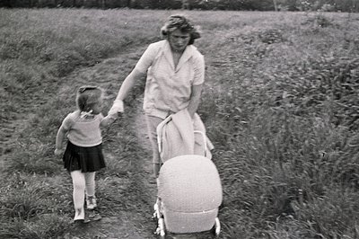 Black & white image: a mother & young girl walk along a path bordered by tall grass. The mother wears a light-colored dress, ...