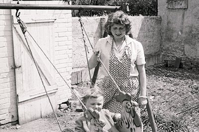 A woman watches a young boy on a whimsical, painted wooden tiger swing. Mid-century backyard scene shows a brick wall and ove...