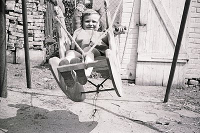 A young child sits on a wooden seat swing, mid-motion, against a backdrop of weathered stone and a clapboard wall. The swing’...