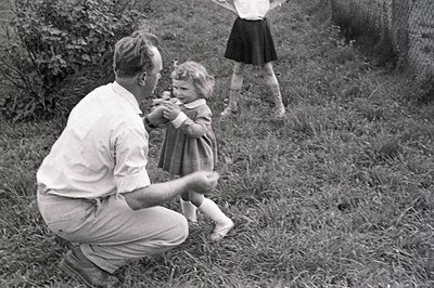 A man in trousers and shirt kneels playfully with a young girl in a dress and socks amidst tall grass. A second child stands ...