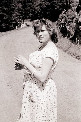 Woman in a floral print dress stands on a paved road, smiling slightly. She wears a bracelet and necklace. Appears to be a ca...
