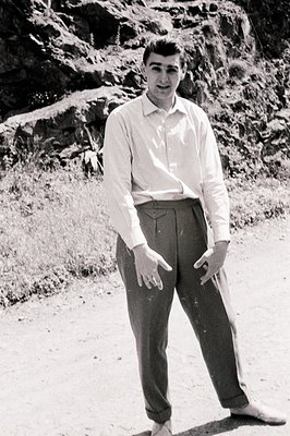 A young man in a crisp, collared shirt and wide-legged trousers stands outdoors, seemingly on a rocky path. He gestures with ...