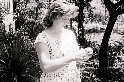 Black and white photo shows a woman in a floral patterned dress, examining an object in her hands. Set in a garden with lush ...