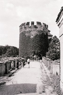 A walled courtyard path leads towards a weathered, ivy-covered stone tower. Several figures, including children, are visible ...
