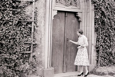A woman in a patterned skirt suit reaches for a wrought-iron door handle within a stone archway, ivy-covered wall to her left...