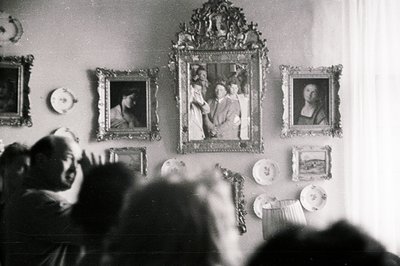 Framed portraits & decorative plates adorn a wall, visible through a blurred foreground of onlookers. Formal portraiture, orn...