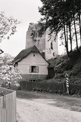 A modest, gabled house sits before the ruins of a medieval stone tower, set within a sloped, wooded landscape. A weathered wo...