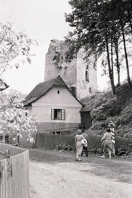Rustic village scene: three figures stroll along a dirt road past a traditional, tiled-roof house adjacent to the ruins of a ...