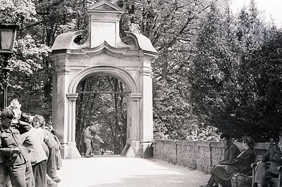 Monochrome image of a monumental stone archway with figures on a pathway & stone retaining wall. Several formally dressed ind...
