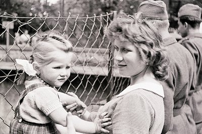 A young girl in a plaid dress and ribboned hair reaches through a chain-link fence towards a smiling woman. Two uniformed sol...