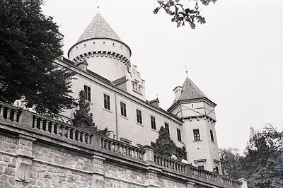 A view of a stone castle with multiple turrets and a balcony, partially obscured by foliage. Two figures are visible on the b...